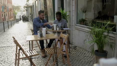 Young-men-using-laptop-and-discussing-work-in-outdoor-cafe